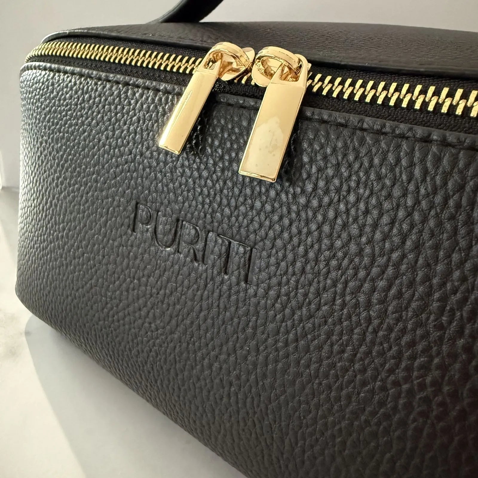 PURITI Medium sized beauty case close-up detail of black pebbled leather and gold-tone zipper pulls and the brand name PURITI embossed on the front.
