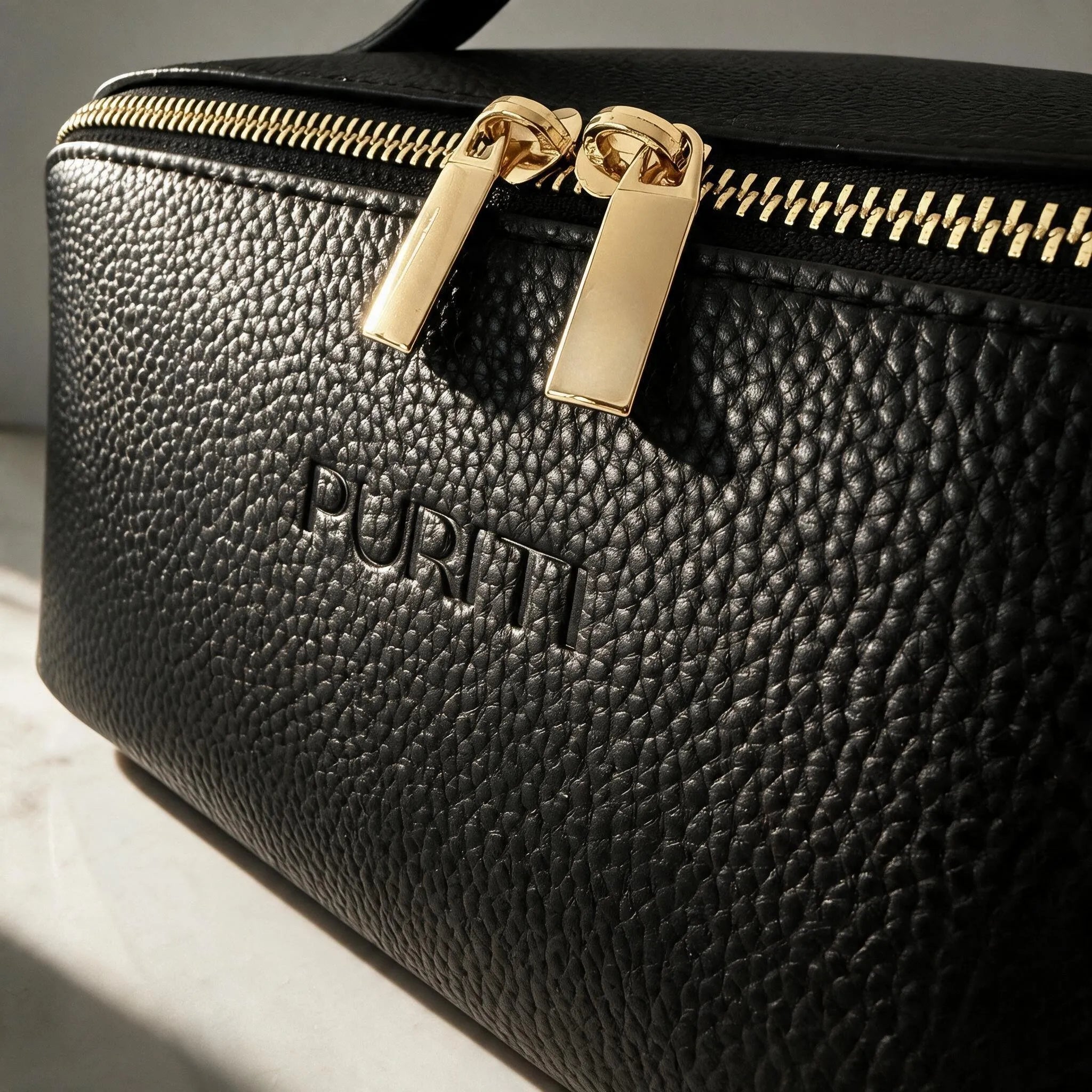PURITI Beauty Case - Medium sized black pebble leather beauty case with gold zipper on a light gray background
