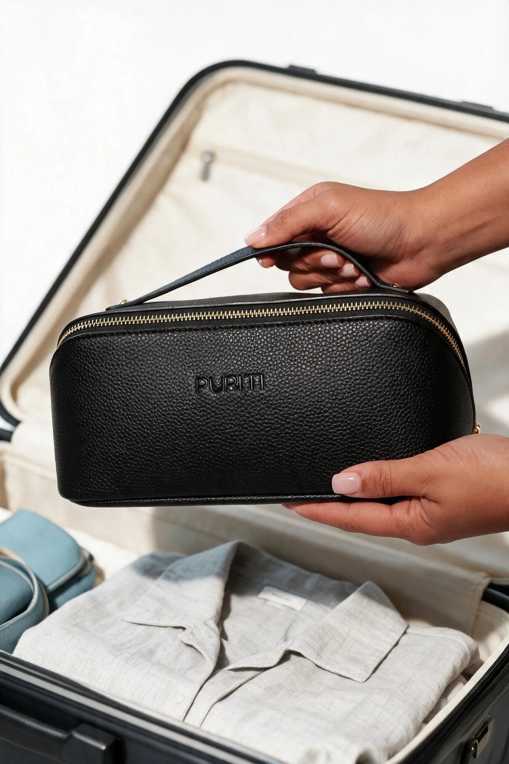 A hand holding the black PURITI Beauty Travel Case by its top handle above a packed suitcase with a folded linen shirt.